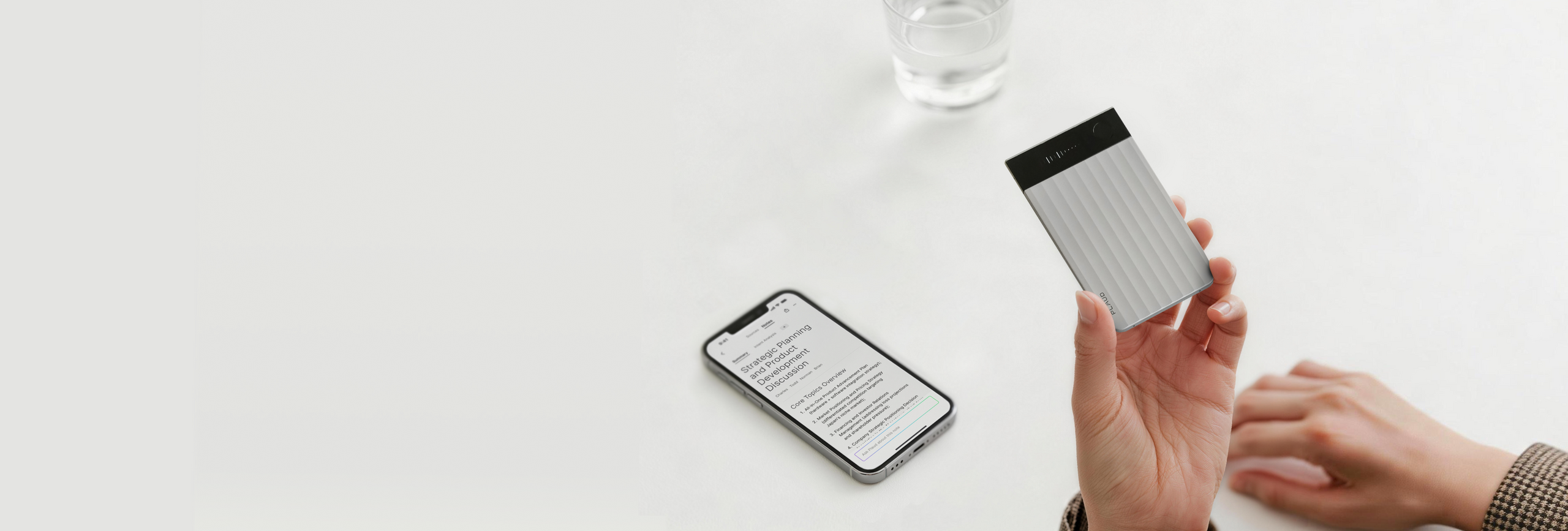 Hand holding a silver Plaud Note Pro with a black top, next to a smartphone displaying text on a white table.