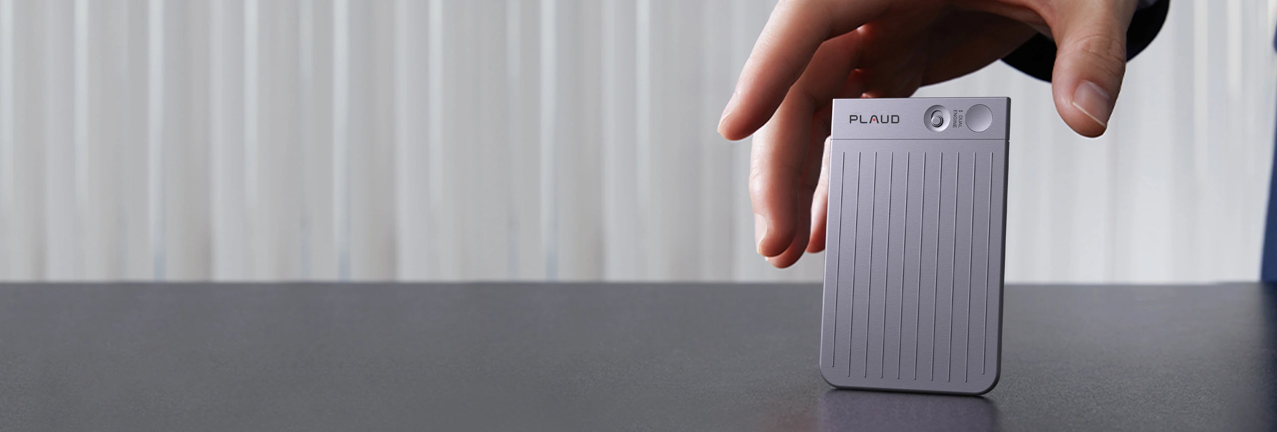 Hand reaching for a silver Plaud Note with ribbed texture, standing upright on a dark table.