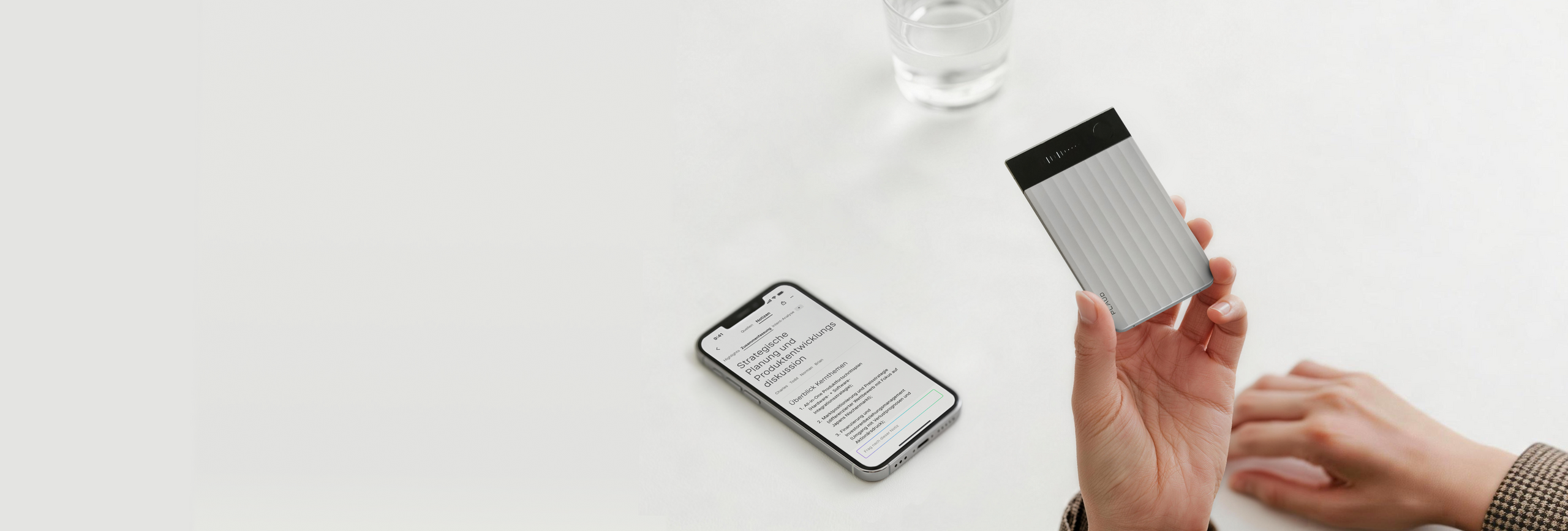 Hand hält silbernen Plaud Note Pro neben Smartphone mit geöffneter Plaud App auf einem weißen Tisch.
