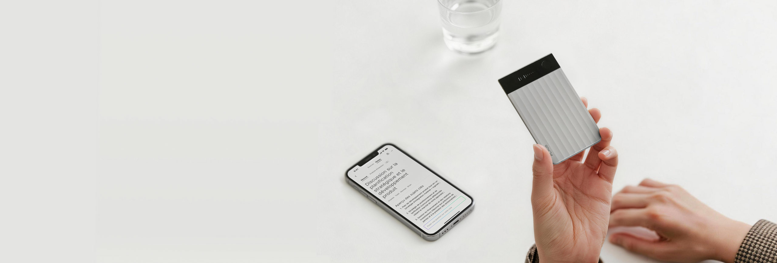 Une main tient un Plaud Note Pro argenté près d'un smartphone affichant du texte sur une table blanche avec un verre d'eau.