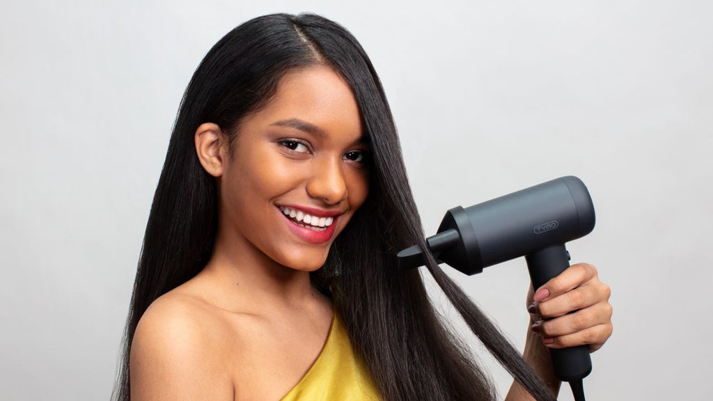 Woman with a radiant smile holding a black hair dryer, styling her sleek, straight hair at home.