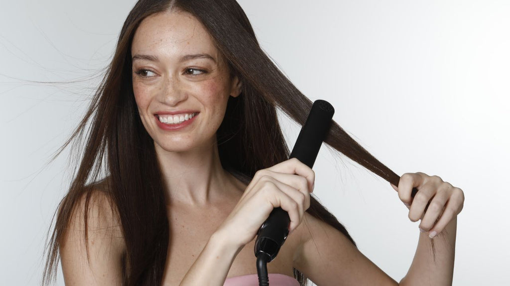 Cheerful woman straightening her hair with a black flat iron, enjoying the ease of styling at home.
