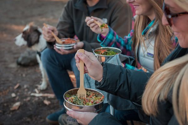 一群人坐在外面,吃一顿饭从不锈钢食品容器使用竹叉。一只狗在背景。