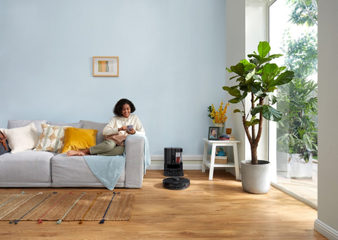 Une jeune fille assise sur le canapé utilise son téléphone portable pour contrôler l'aspirateur robotisé Roborock (S7 MaxV Ultra) dans sa maison afin de nettoyer les acariens du sol.