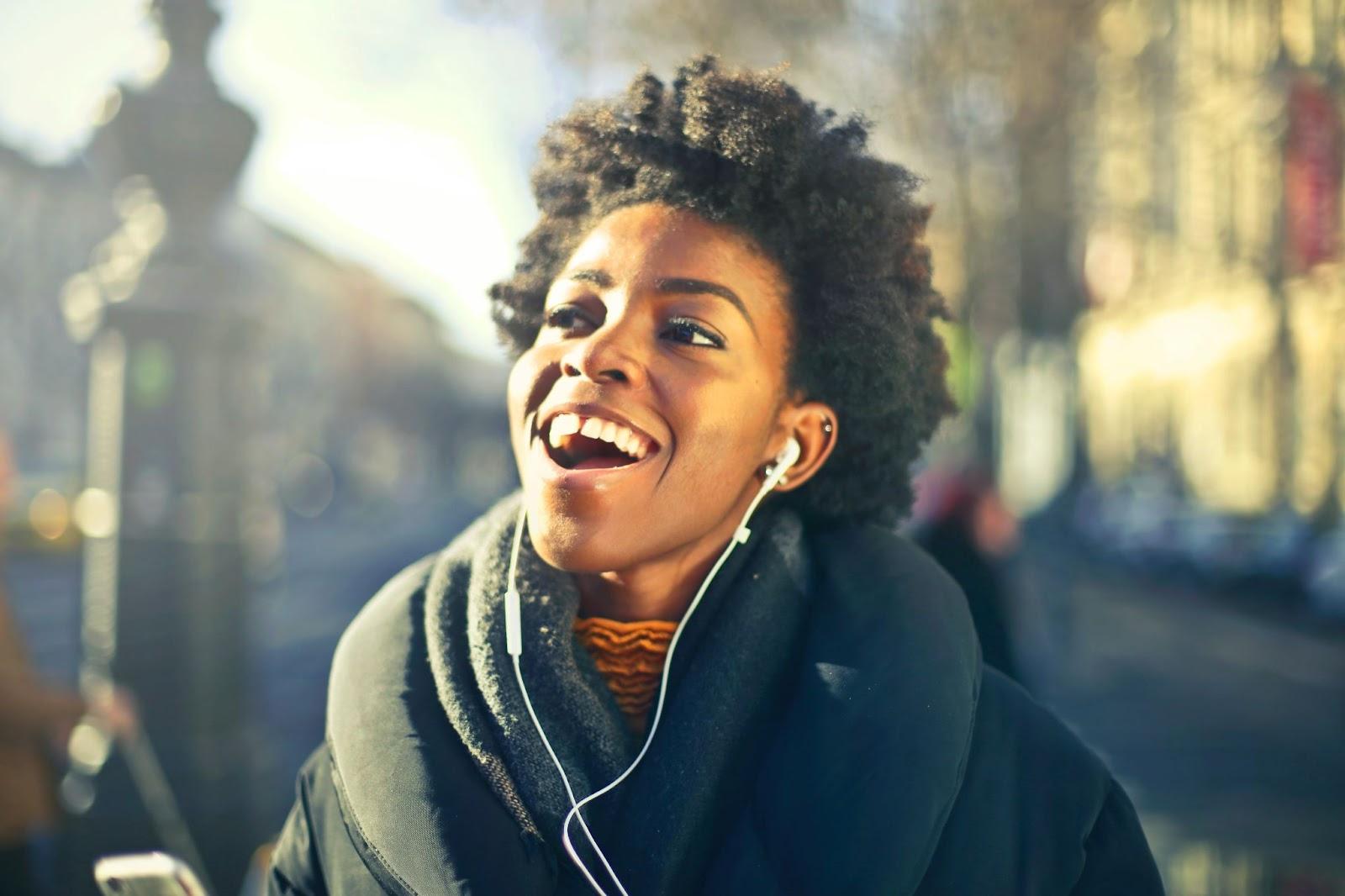  A person wearing wireless headphones with a built-in microphone, listening to music on their mobile phone. The headphones are sleek and stylish, with a Bluetooth logo visible on the side. The person is smiling and immersed in their music experience