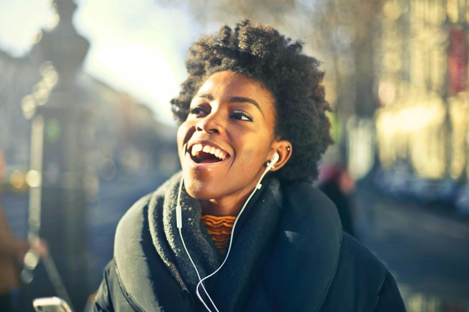 A person wearing wireless headphones with a built-in microphone, listening to music on their mobile phone. The headphones are sleek and stylish, with a Bluetooth logo visible on the side. The person is smiling and immersed in their music experience