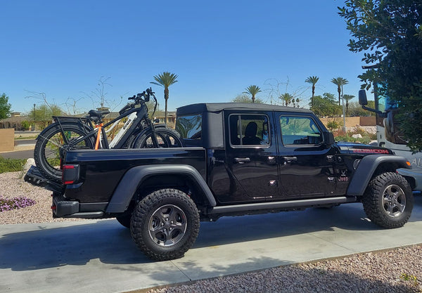 Black Jeep Gladiator parked on driveway with two bicycles mounted on the back, palm trees and clear sky in the background.