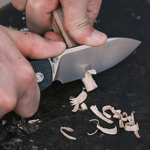 A Vosteed Raccoon pocket knife with a black textured handle is shown whittling wood, highlighting its sharp blade