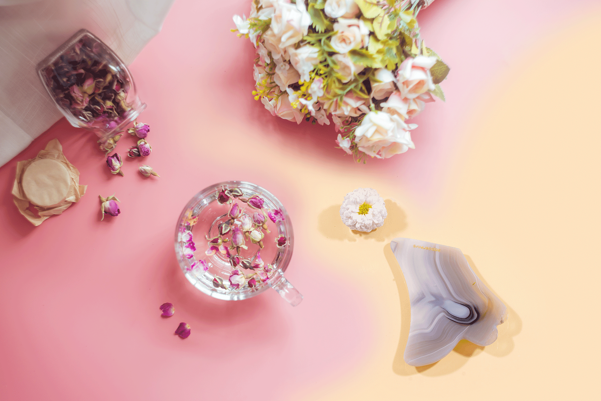 a cup of flower tea and rose flower lay flat on a pink bacnground as well as a piece of agate gua sha stone