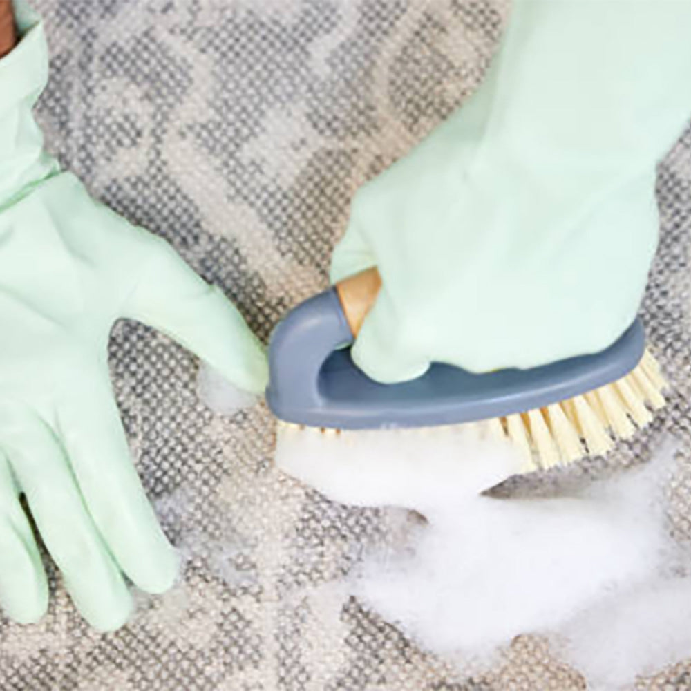On the grey carpet, a man with light green leather gloves. He is brushing the carpet with a blue plastic brush. White foam appeared on the carpet.