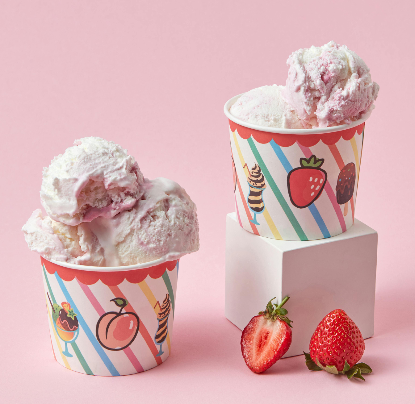 Two scoops of strawberry ice cream in colorful Paper Ice Cream Cups, with fresh strawberries on a pink background.
