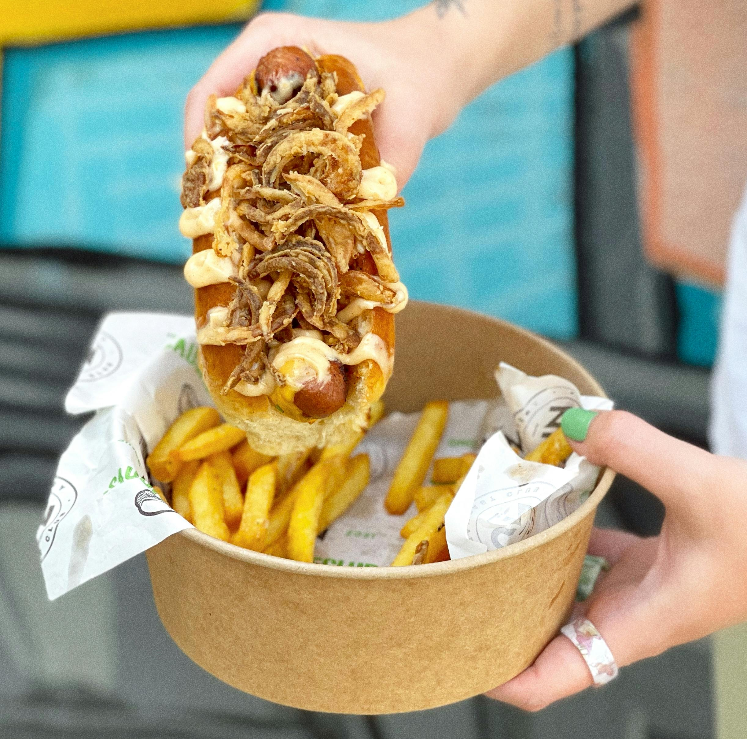 Hands holding a hot dog with fried onions and sauce, and crispy fries in a patterned paper bowl; classic fast food combo.