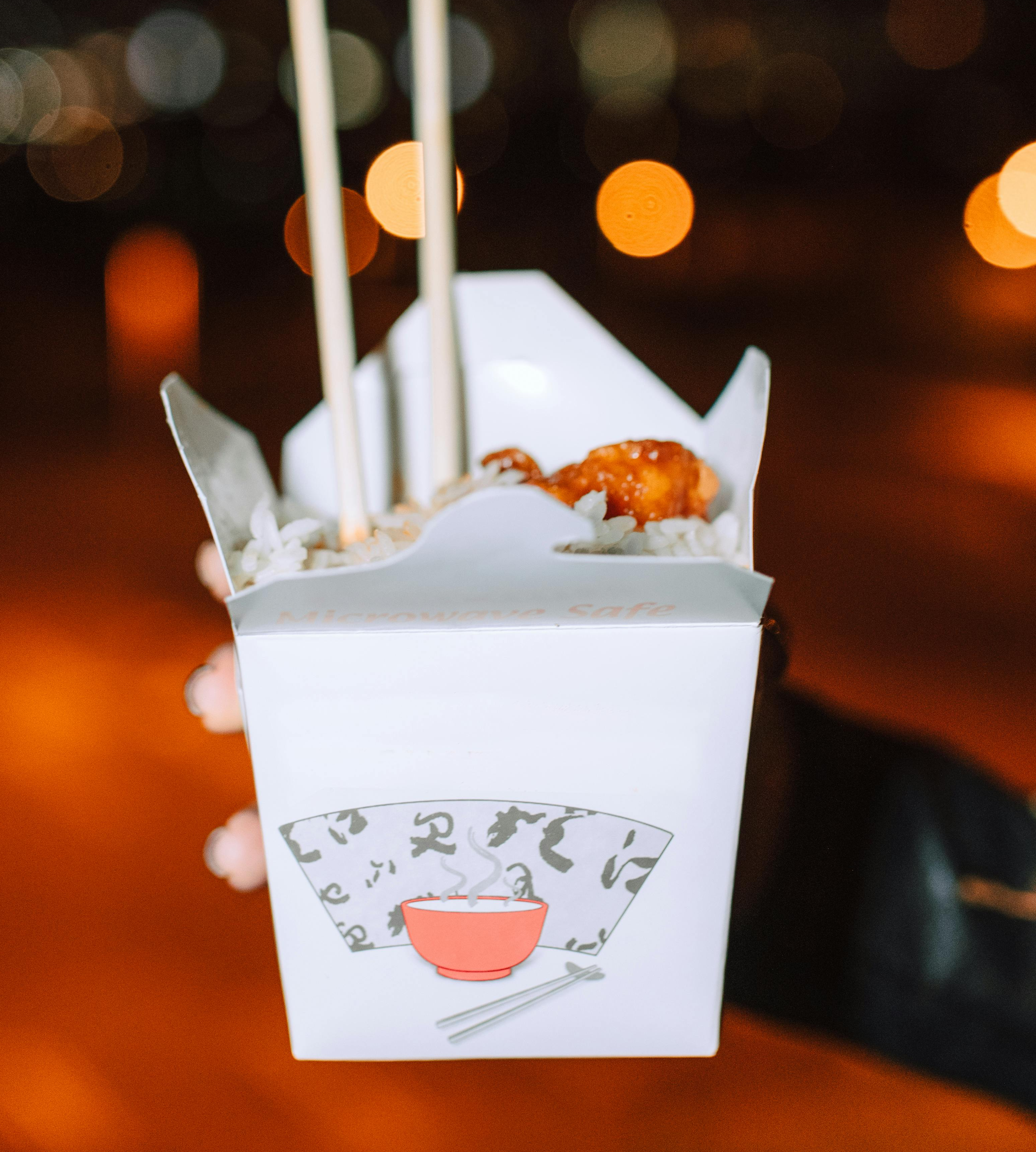 A hand holds a white paper box with Japanese-inspired design, filled with rice, saucy chicken, and chopsticks.