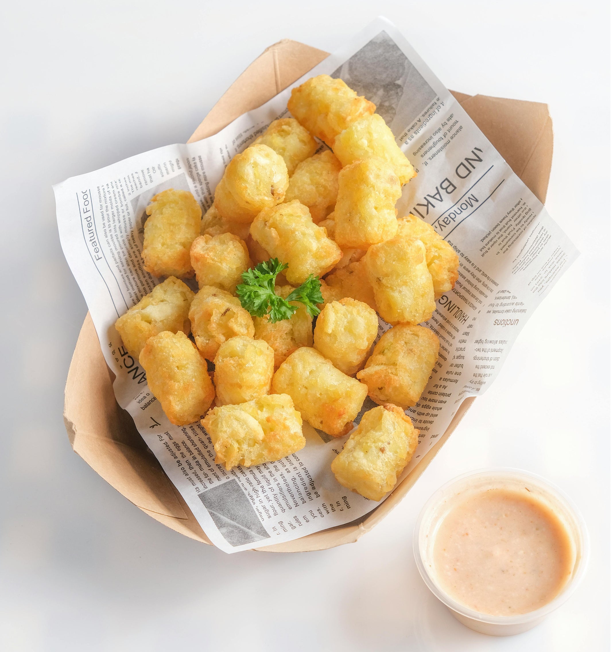 Crispy golden fries in a brown paper box lined with newspaper, garnished with parsley, and served with a small cup of orange dipping sauce.