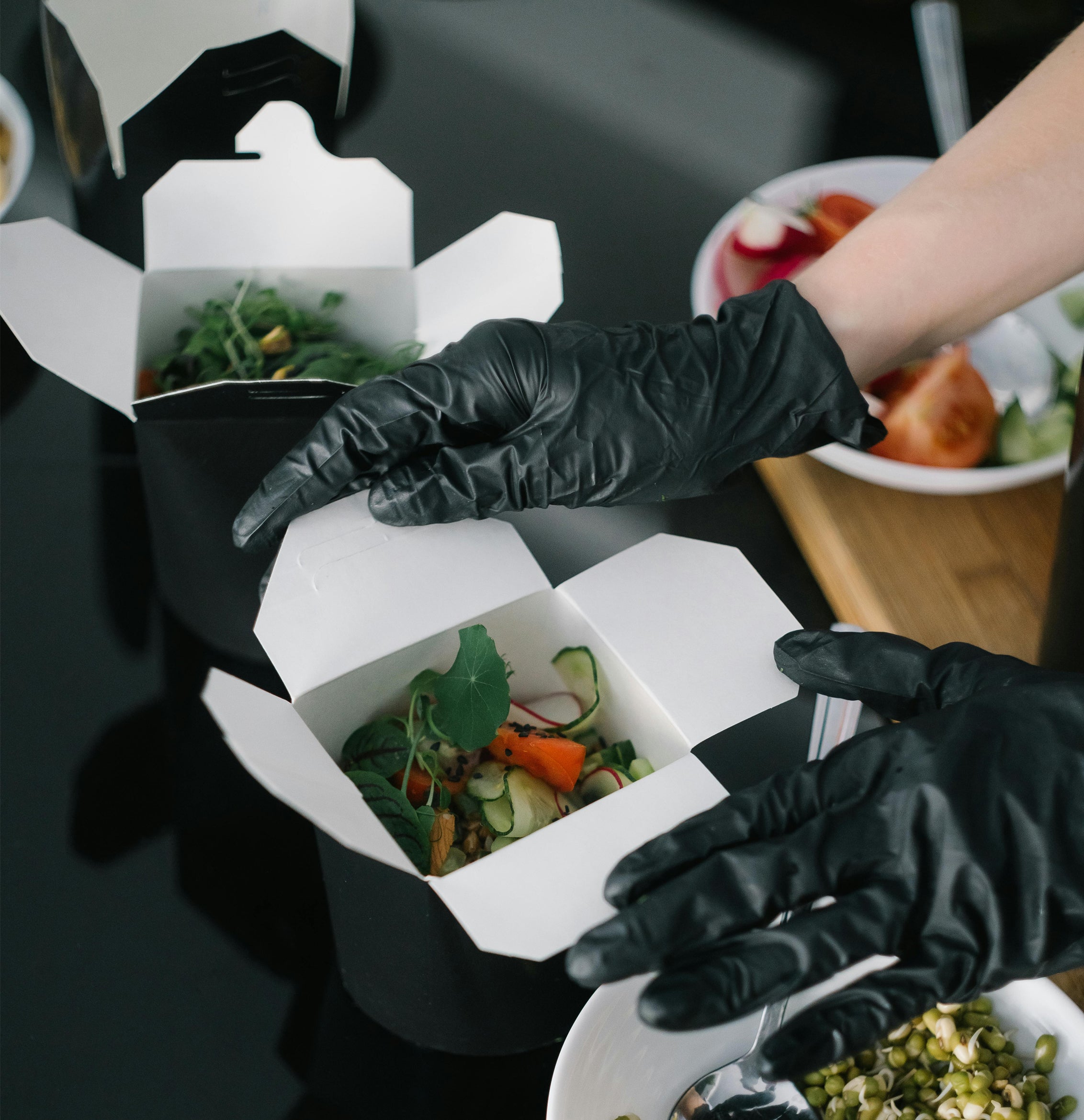 Hands wearing black gloves handling black and white paper boxes filled with colorful vegetable salad.