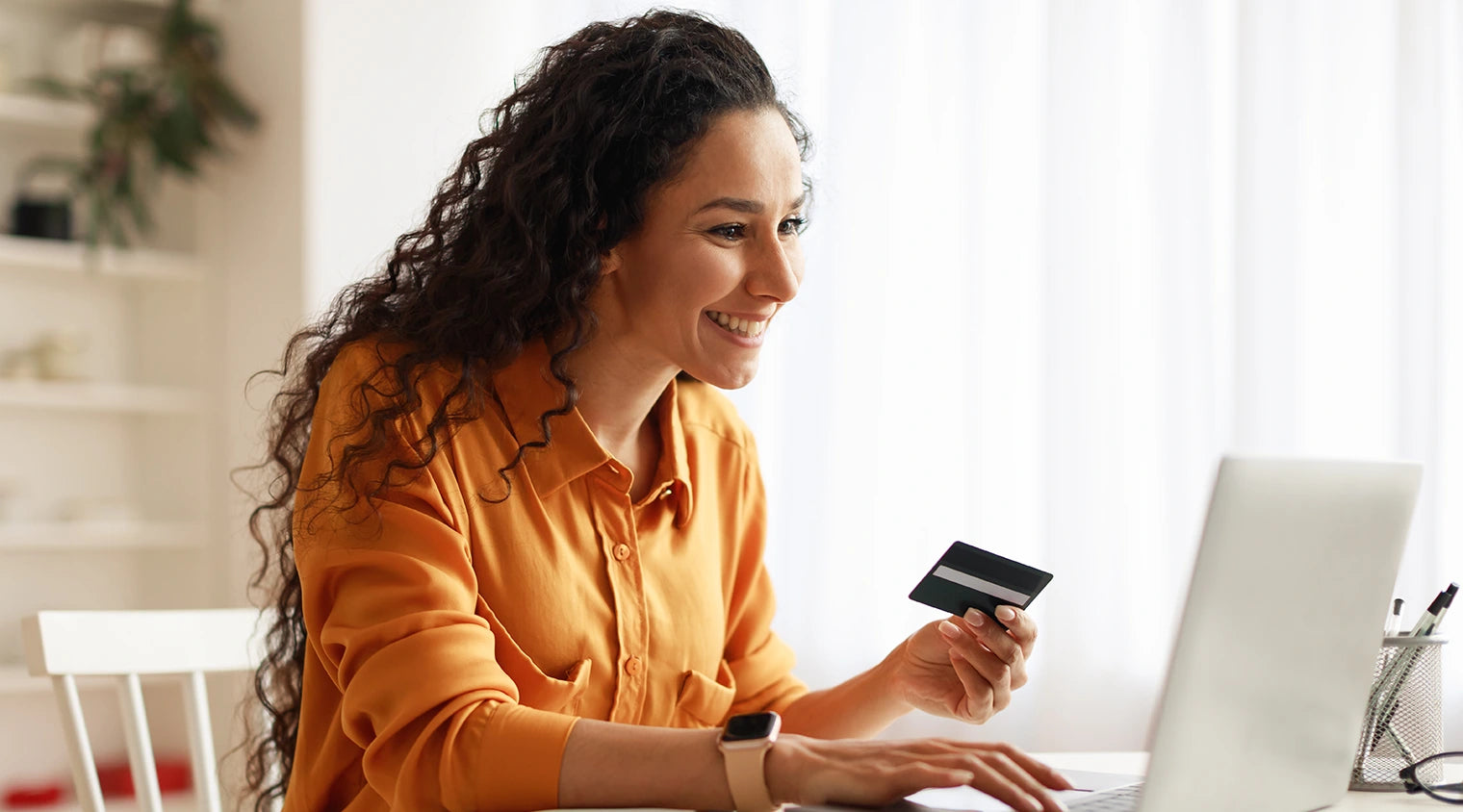 A smiling woman with curly hair sits at a white table, holding a credit card in her right hand and typing on an Empava laptop with her left hand. She wears an orange shirt and a smartwatch. A white curtain and a plant in the background provide a bright setting.
