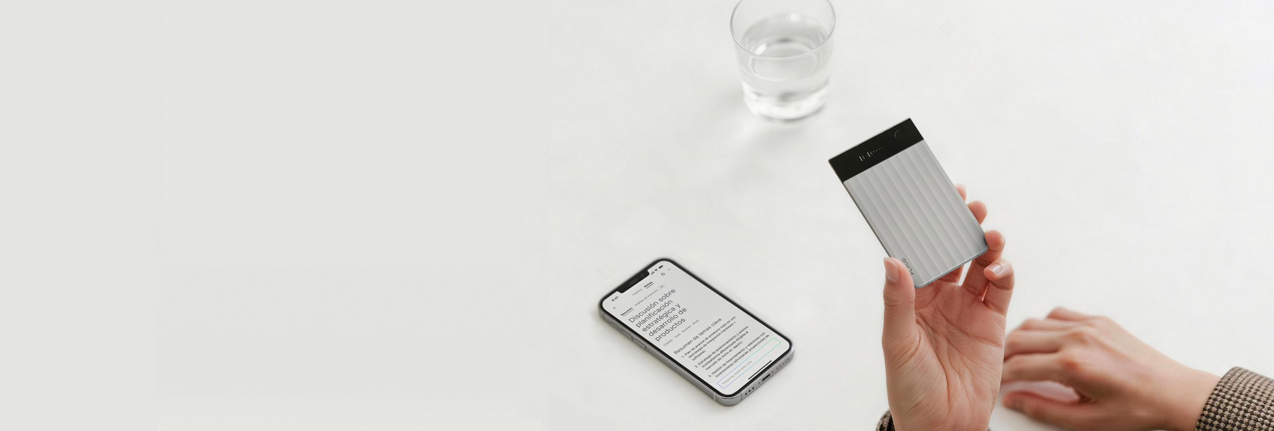 Mano sosteniendo un Plaud Note Pro plateado con pantalla, junto a un smartphone mostrando texto en una mesa blanca.