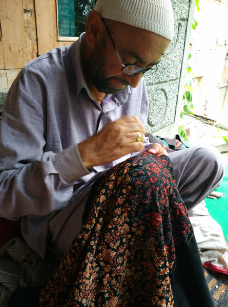 Un maestro ricamatore sta cucendo a mano uno scialle di pashmina, seduto fuori alla luce naturale del giorno, con lo scialle appoggiato sulle ginocchia.