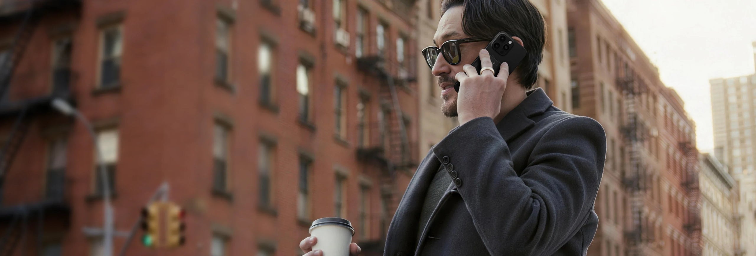 A man in a dark coat talks on a black smartphone with a black Plaud NotePin attached, holding a coffee cup on a city street.