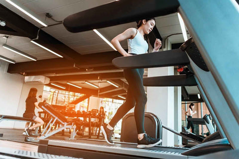 A woman dressed in sports attire is exercising on a treadmill inside a gym. Her hair is blowing in the wind, indicating her focus and energy. She wears black leggings, a white tank top, and gray sneakers. In the background, other gym equipment is visible, and blurred figures of individuals can be seen engaging in various activities.