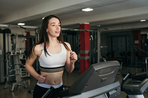 A photograph of a young woman exercising on a treadmill in a gym. She wears a white tank top and black sports shorts, with her hair flowing freely. Her expression is focused and determined as she appears to be running vigorously. In the background, other fitness equipment such as dumbbell racks and machines can be seen.