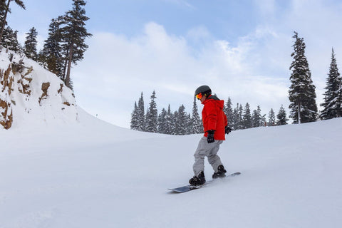 A person wearing a red jacket and gray pants is skiing on a snow-covered hillside. They wear a black helmet and orange goggles, and appear to be enjoying the thrill of winter sports. The background consists of snowy slopes, trees, and a few clouds in the sky.