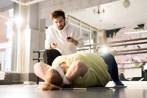 A man wearing a white sweater and black pants stands next to a woman lying on the ground in a gym. He stands to her right and holds a remote control in his hand, seemingly operating a fitness equipment or monitoring system. The woman wears a yellow top and gray sports pants, with white headphones on her head. She lies on her back with her hands by her sides and her legs stretched out.