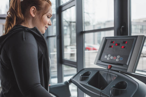 A woman wearing a black sports outfit with her hair tied in a ponytail using a treadmill in a gym. She stands side-on, focusing intently on the display screen of the treadmill. The treadmill is positioned next to a large, bright window, allowing a view of buildings and vehicles outside. The treadmill's display shows 00:00 for time, 0 km for distance, 0 km/h for speed, and 0% for incline. Other fitness equipment and machines are visible in the background.