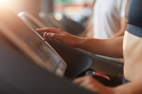 A hand wearing light-colored clothing operates the touchscreen of a treadmill. The treadmill is situated in an indoor environment, with a blurred background that faintly reveals other people and fitness equipment. The hand gestures appear to be adjusting the speed or settings of the treadmill. Sunlight streams in from the left side of the frame, adding a warm ambiance to the scene.