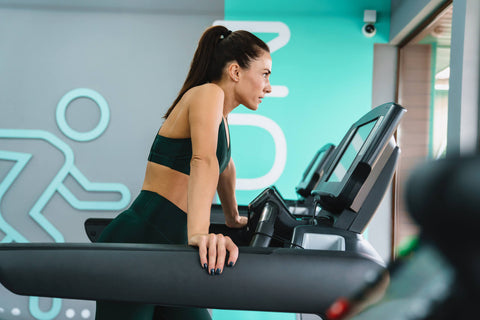 A woman is using a treadmill in a gym. She wears a green sports vest and tight pants, with her hair tied in a ponytail. She is focused and looking straight ahead. The treadmill is black with a display screen and control panel. In the background, there is a wall with a white human figure drawn on it, with the letter 'P' beside it.