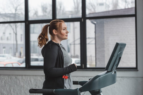 A woman exercising on a treadmill in a gym. She wears a black sports top and gray pants, with her hair tied in a ponytail. She focuses ahead with a determined expression. The treadmill is situated indoors, with a window view of parked cars outside.