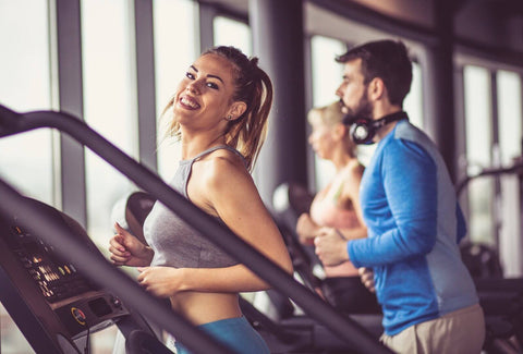 A group of people exercising on treadmills in a gym. In the foreground, a blonde woman wearing a gray tank top and blue exercise pants smiles happily while running on a treadmill. Her hair is tied in a ponytail, and she leans slightly forward with both hands gripping the treadmill handles.