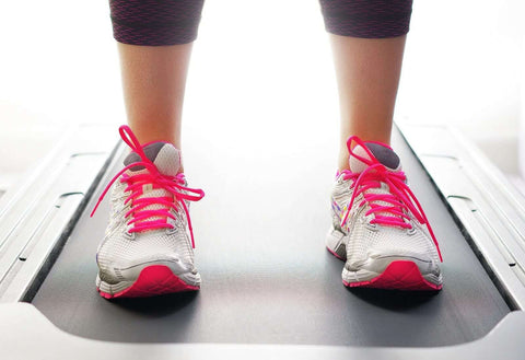 This image shows a person standing on a treadmill wearing pink-accented white sneakers with laces. The treadmill is black, and the background is blurred, obscuring specific details. The lower half of the person's body is cut off, leaving only the gray leggings and ankles visible.