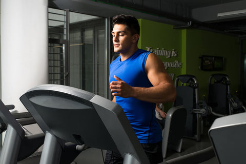 A young man dressed in a blue sports vest and black shorts exercising on a treadmill inside a gym. His facial expression is focused, indicating that he is engaged in a cardio workout. The treadmill is positioned in a bright room with green walls and a banner hanging on one side reading 'Training is possible' (correction of the apparent typo 'ainingis pposite' on the banner). Other fitness equipment such as dumbbell racks and bicycles are visible in the room.