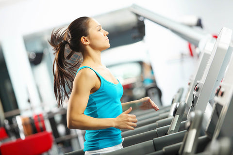 A photograph of a young woman exercising on a treadmill in a gym. She wears a blue tank top and gray sports pants, with her hair tied in a ponytail. She focuses intently ahead as she runs. The treadmill is located indoors, and in the background, other fitness equipment and blurred figures can be seen.