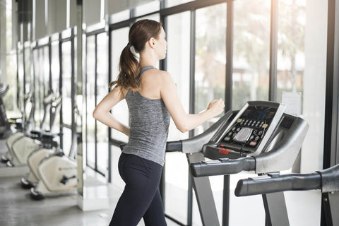 An image showing a woman using a treadmill in a gym. She wears a gray vest and black leggings, with her hair tied in a ponytail, focused ahead. The treadmill is located near a row of windows, where trees can be seen outside. Other fitness equipment such as dumbbell racks and bicycles are visible in the background. The entire scene conveys a sense of serenity and organization.