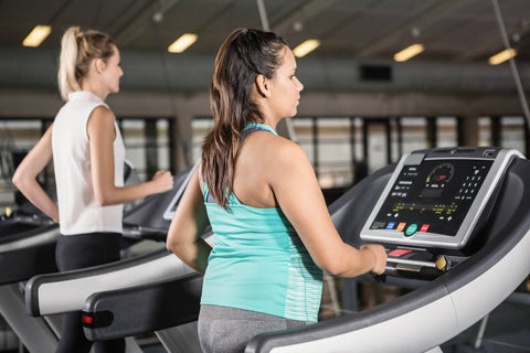 A woman in a pink tank top and gray leggings, wearing a pink baseball cap, is actively exercising on a treadmill in a gym. The gym interior is visible in the background, featuring large windows that allow natural light to flood the space, along with other fitness equipment.