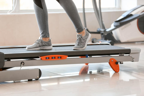 A person dressed in gray sweatpants and white sneakers is standing on a black treadmill with orange accents, preparing for a workout. The treadmill is located in a bright indoor environment, with a blurry fitness machine visible in the background.