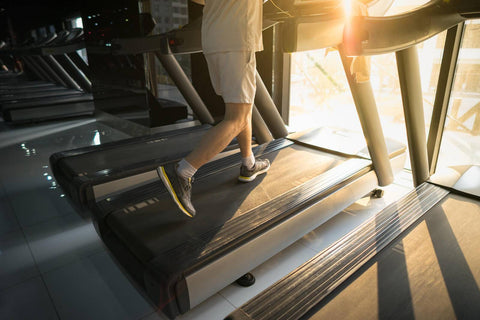 A person wearing white shorts and gray sneakers is actively exercising on a treadmill in a gym. Sunlight streams through the windows, creating bright spots of light. The treadmill stands in an indoor setting, and several other treadmills can be seen in the background. The individual has a focused expression, alternating their feet as they walk forward with a slight forward lean, indicating an intense cardio workout.