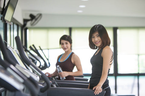 A photograph of two women using a treadmill in a gym. They stand in front of a black treadmill, with windows and other fitness equipment visible in the background. The woman on the left wears a brown top and dark pants, while the woman on the right is dressed in a black sleeveless top and black pants. Both women smile brightly, appearing in good spirits.