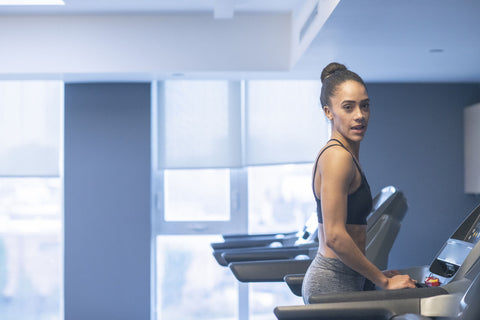 A photograph showcasing a woman utilizing a treadmill in a gym. She wears a black sports tank top and gray leggings, her hair tied into a high ponytail. Her expression is focused and serious, indicating she is engaged in a rigorous exercise routine.