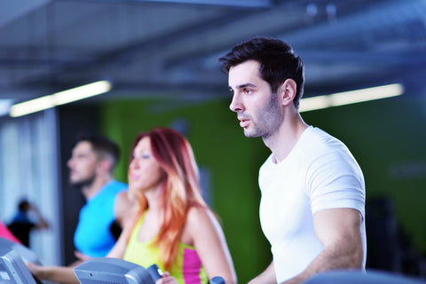 A male individual dressed in a white top and dark pants is actively running on a treadmill in a gym. His gaze is directed towards the left, seemingly focused on something or someone. Behind him, two women are also exercising on separate treadmills, one wearing a yellow top and the other a blue top.