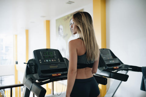 A blonde woman standing on a black treadmill in a bright gym, facing away from the camera. She wears a black sports top and tight shorts. The treadmill is located in a spacious area with yellow walls and windows visible in the background. Other fitness equipment is also seen in the distance.