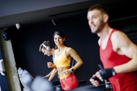 A photo capturing three individuals exercising on treadmills in a gym. On the left, a male wearing gray workout attire and headphones is running actively. In the center, a smiling female dressed in a yellow tank top and red leggings is also jogging. To the right, a male in a red sleeveless shirt and black shorts stands focused on the treadmill ahead. The background reveals the interior of the gym, featuring dark walls and lighting fixtures.
