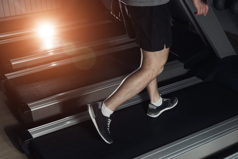 A person wearing black shorts and a gray T-shirt is exercising on a treadmill in a gym. The lower half of the person's body, including their legs and feet, is clearly visible.