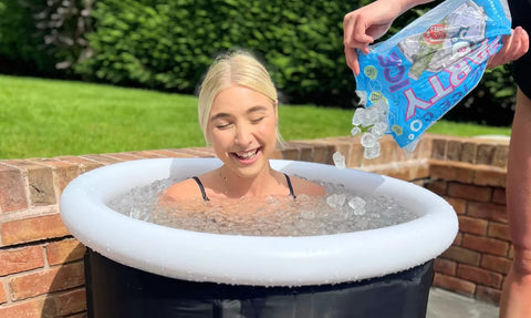 A woman smiles while sitting in an ice bath, with ice being poured into the tub, indicating the refreshing and therapeutic experience of cold plunging.