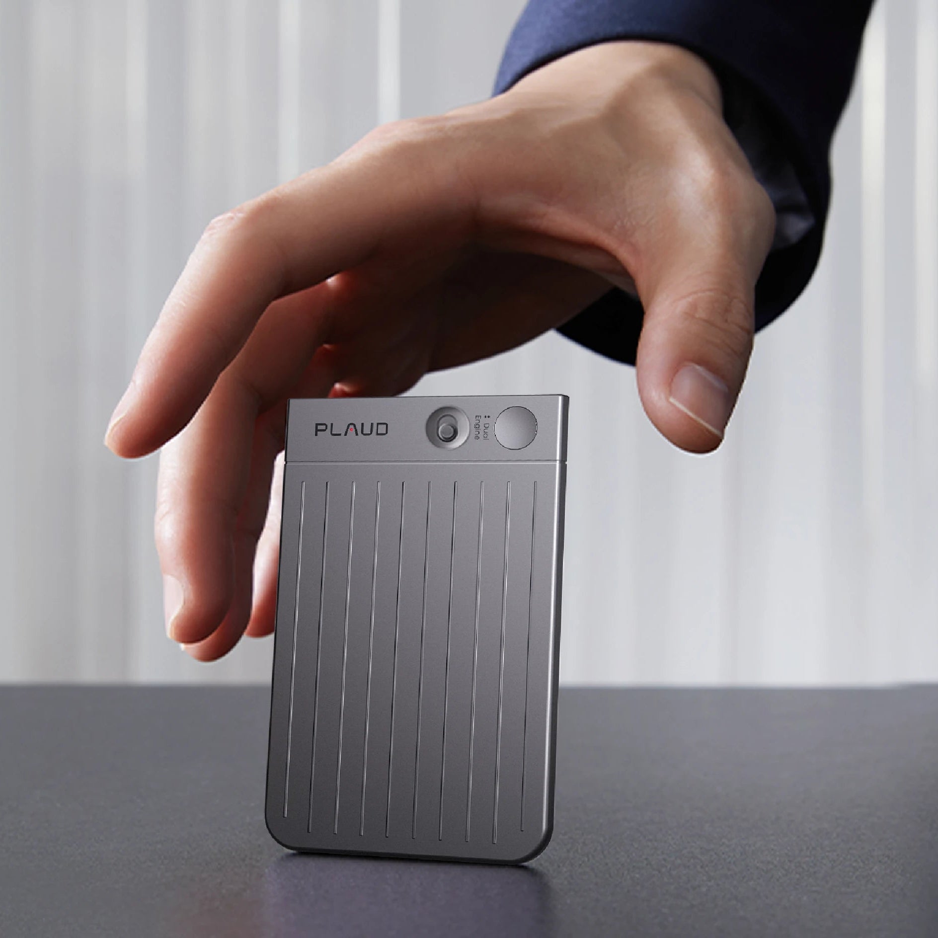 A hand reaching for a gray Plaud Note Pro AI voice recorder standing upright on a dark surface.