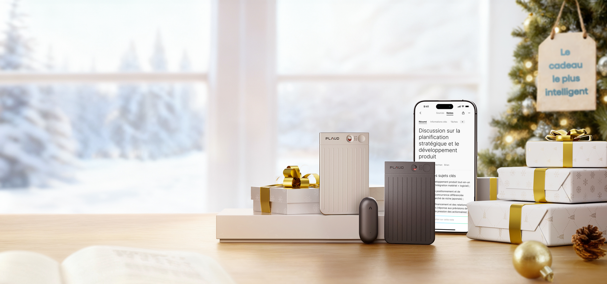 Deux Plaud Note (beige et gris foncé) et un Plaud NotePin gris foncé sur une table en bois, avec des cadeaux et un sapin de N
