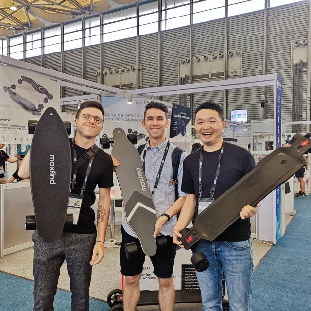 三位男性在展会摊位前各举起一块 maxfind 滑板